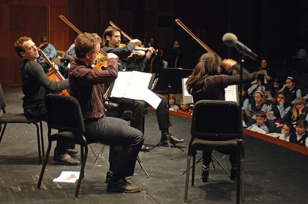 Four violinists perform on an auditorium stage to an audience of elementary school-age children wearing uniforms.