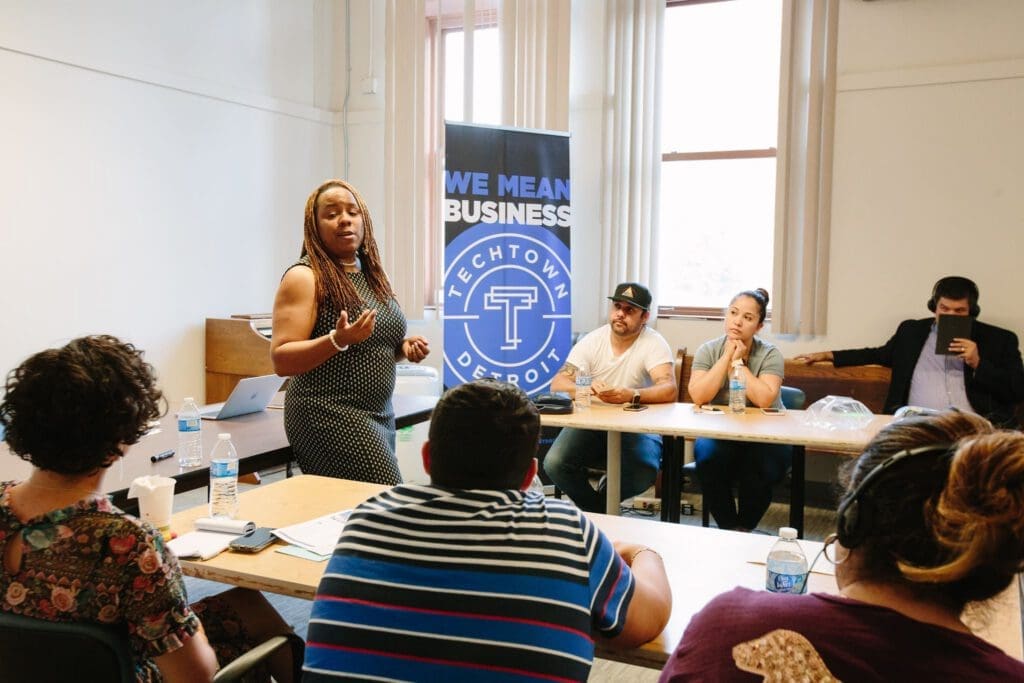 A woman speaks in front of a group of people who are seated behind wooden desks. 
