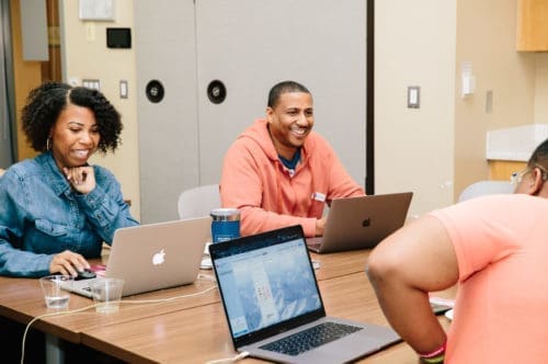 Participants sitting at table working on laptops and smiling at product development day.