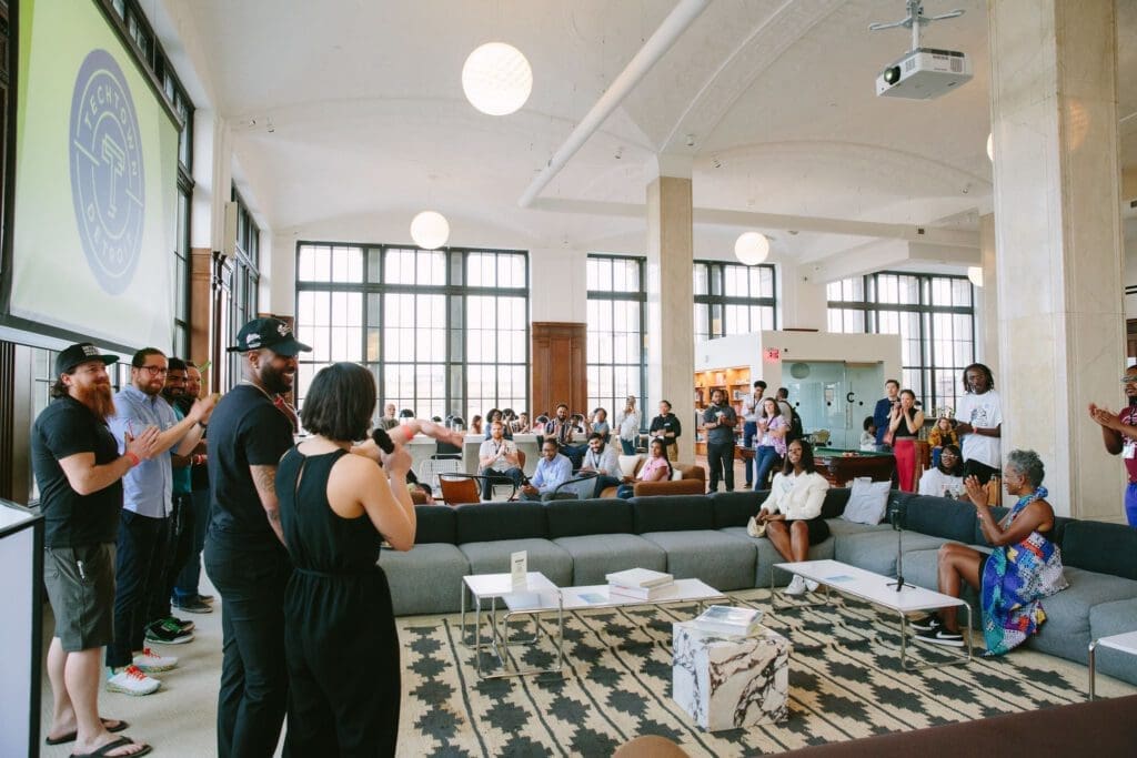 A man smiles next to a woman holding microphone who is speaking to a crowd of people standing and sitting on couches and chairs in a well-lit event space. Behind the man stands four men standing under a projector hanging from the ceiling. The projector shows TechTown Detroit's blue and white logo. 