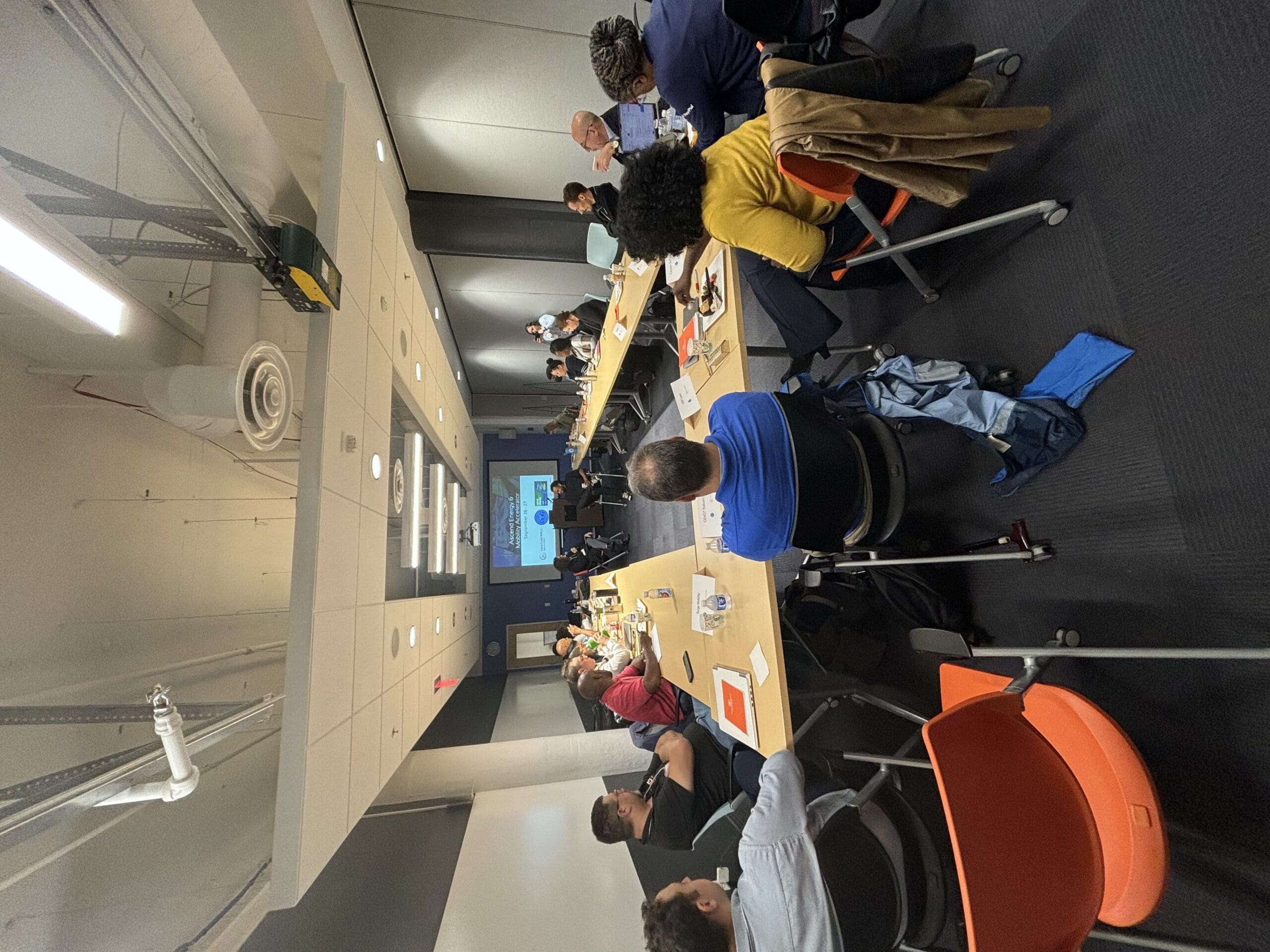A group of people sit at long desks, round-table style, for a workshop held in a conference room. They're all looking at the front of the room, where two people are talking in front of a projector. 