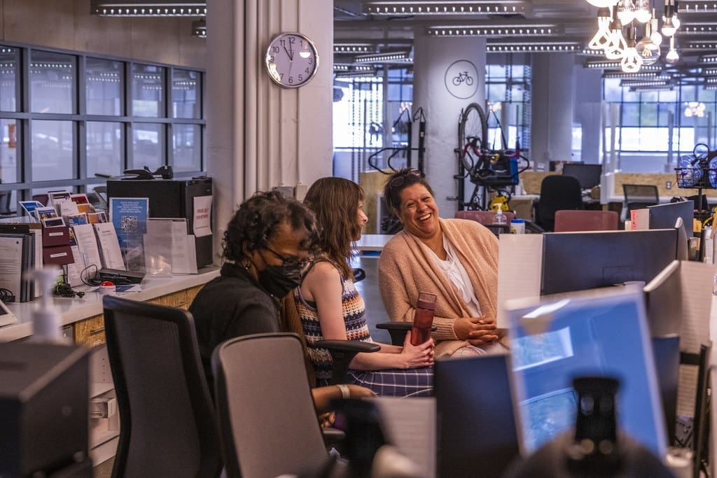 Three women sit and talk behind a front desk.