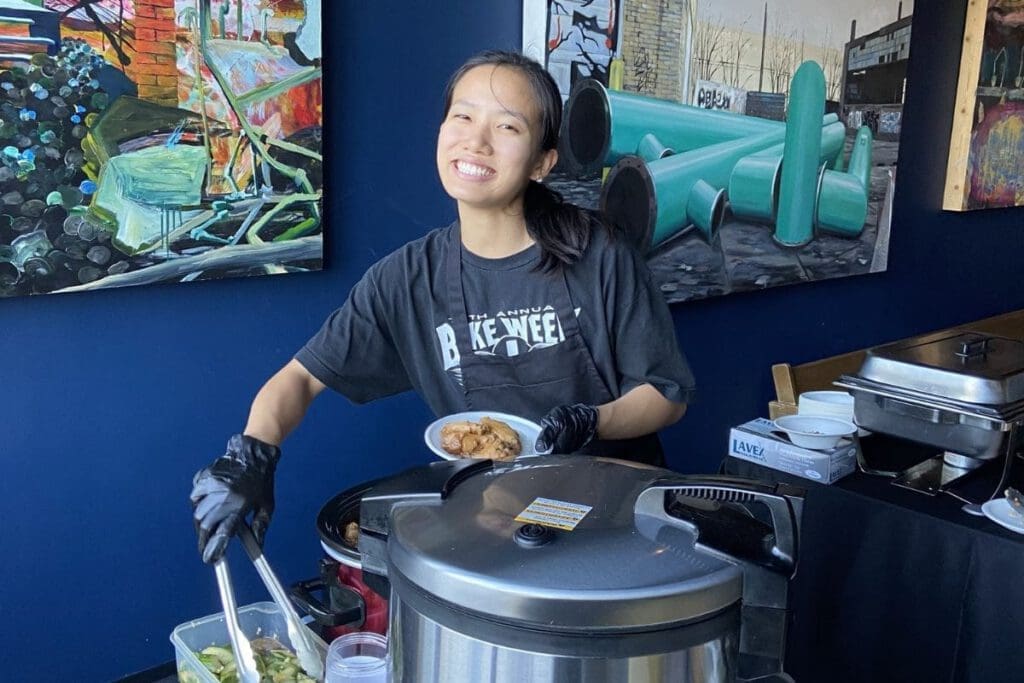 A woman wearing a dark grey shirt, black apron, and black gloves smiles as she prepares food. She holds a bowl of food in her left hand and is using tongs to pick up food with her right hand.