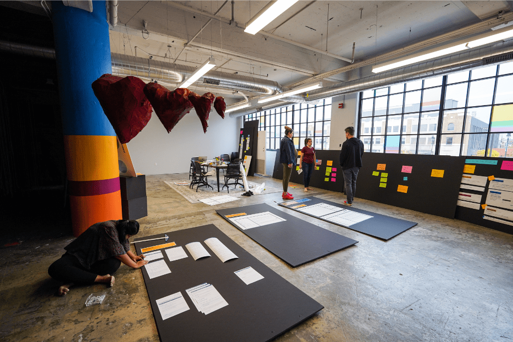 A photo of an open office space. There are large black poster boards with papers and sticky notes on them that people are standing and sitting around.