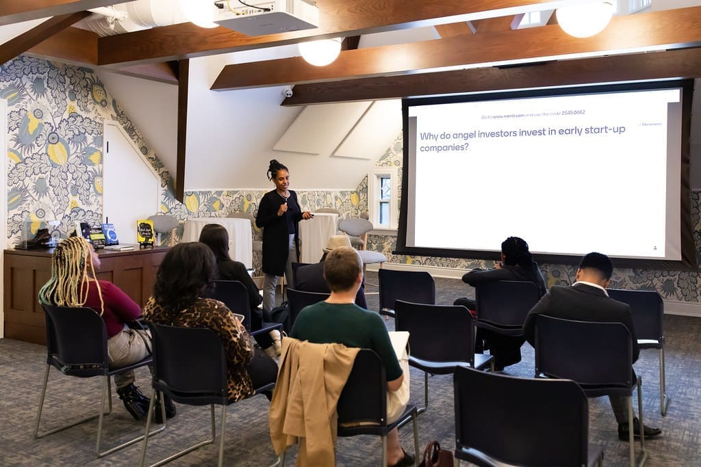 A woman stands to the side of a projector and speaks to a group of six individuals seated throughout three rows of chairs