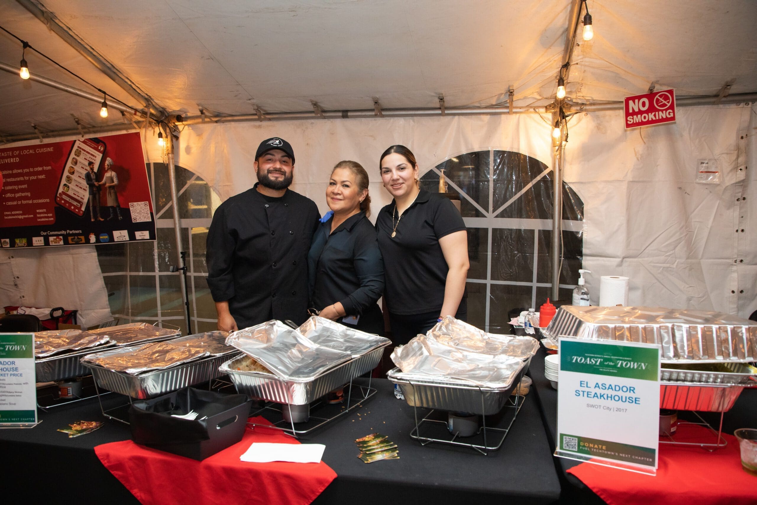 Three people smile as they stand behind a table of food from El Asador Steakhouse in Detroit