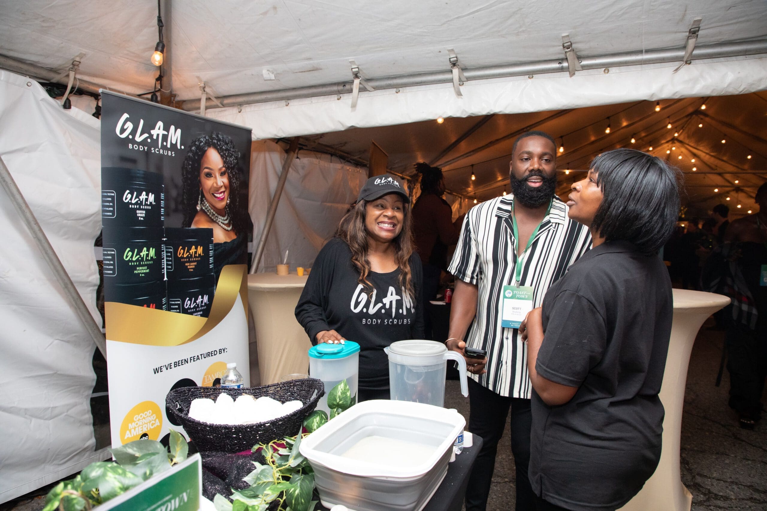 Three people stand and talk to each other, as they stand around a table display for the online skin care business, G.L.A.M. Body Scrubs by Amarra Products