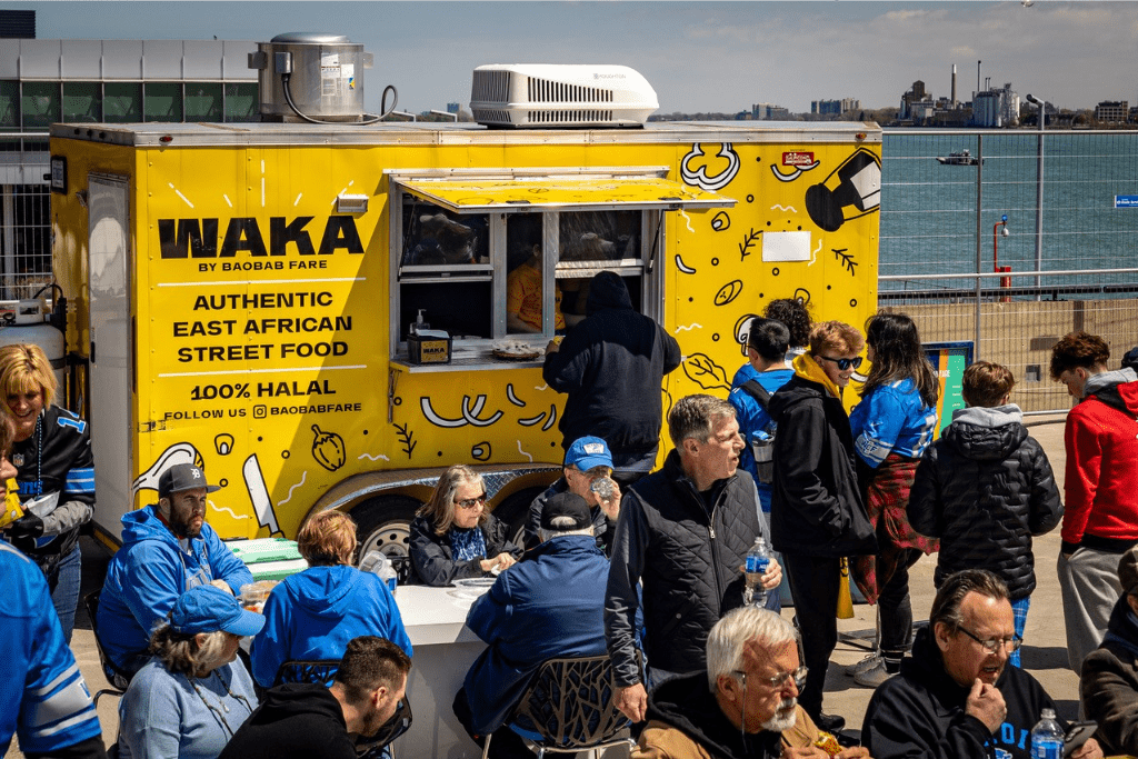 A group of people standing, sitting and eating outdoors, surrounding the Waka by Baobab Fare food truck
