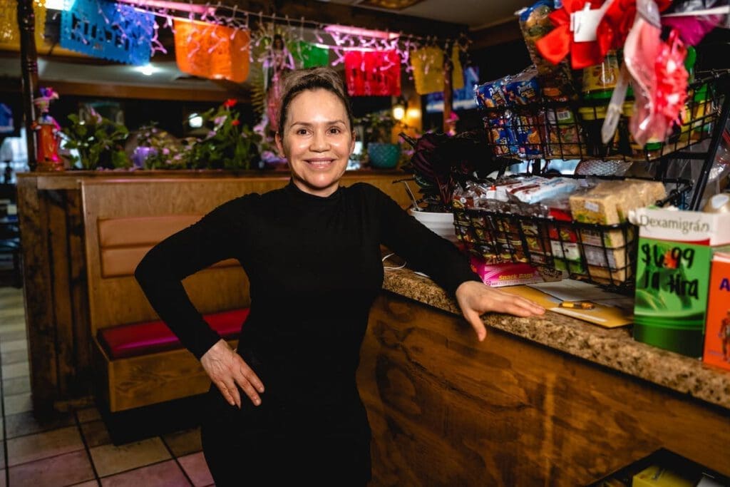 A woman smiles and poses for a photograph, with her right hand on her hip and her left hand resting on a table. 