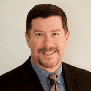 A man smiling for a headshot, wearing a black suit jacket, grey-blue dress shirt, and brown and gray tie