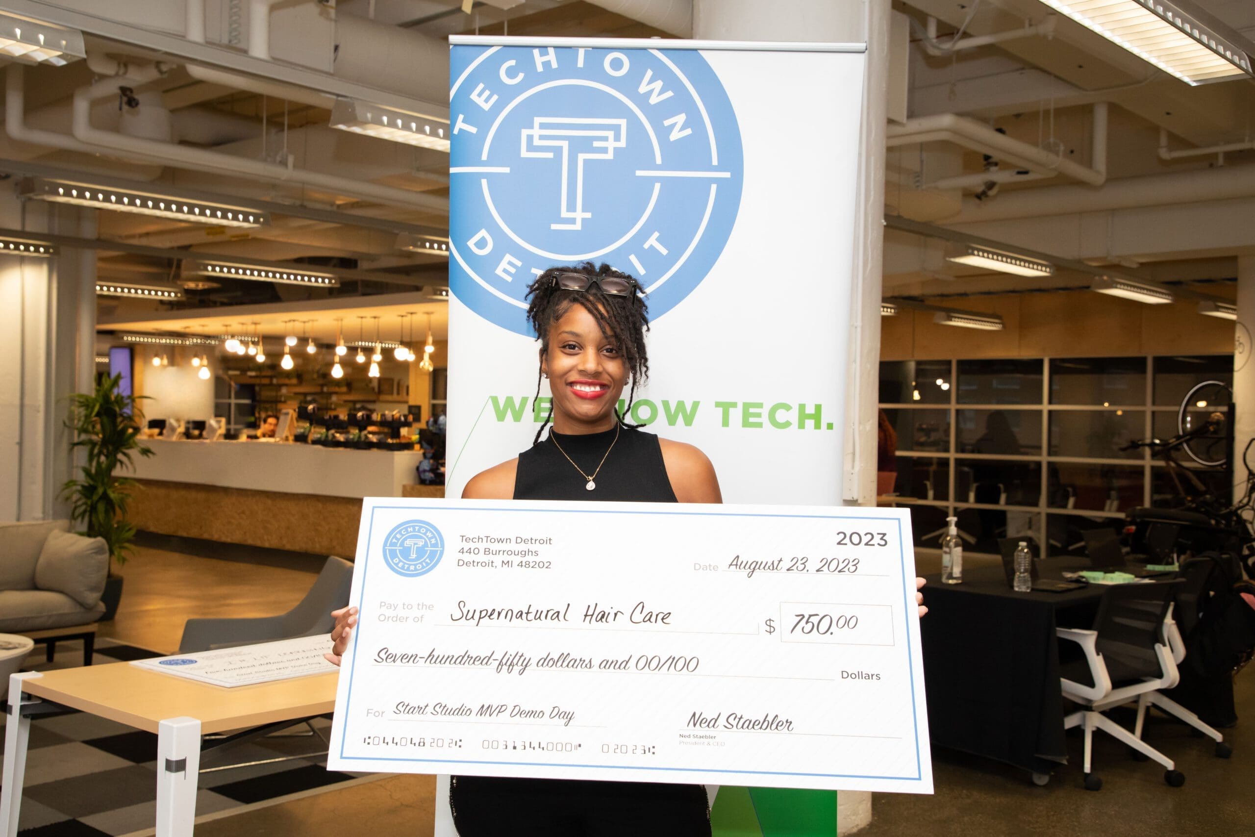 A woman of color holds a large check for $750, for her business, Supernatural Hair Care