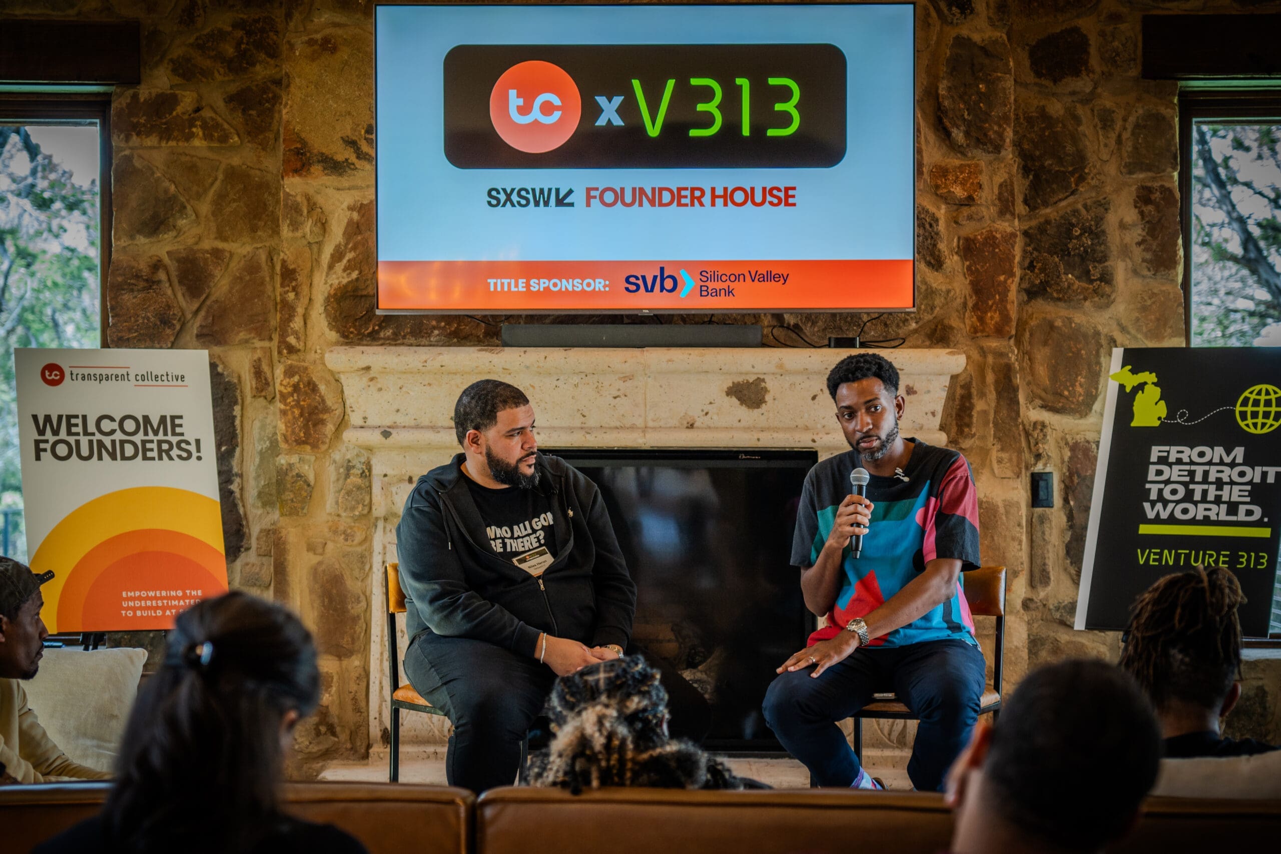 Two people sitting in front of a fireplace, speaking to people sitting in front of them on a couch. There are two posters and a TV screen depicting promotion for the event, TC x V313 @ SXSW: Program for Underrepresented Founders 