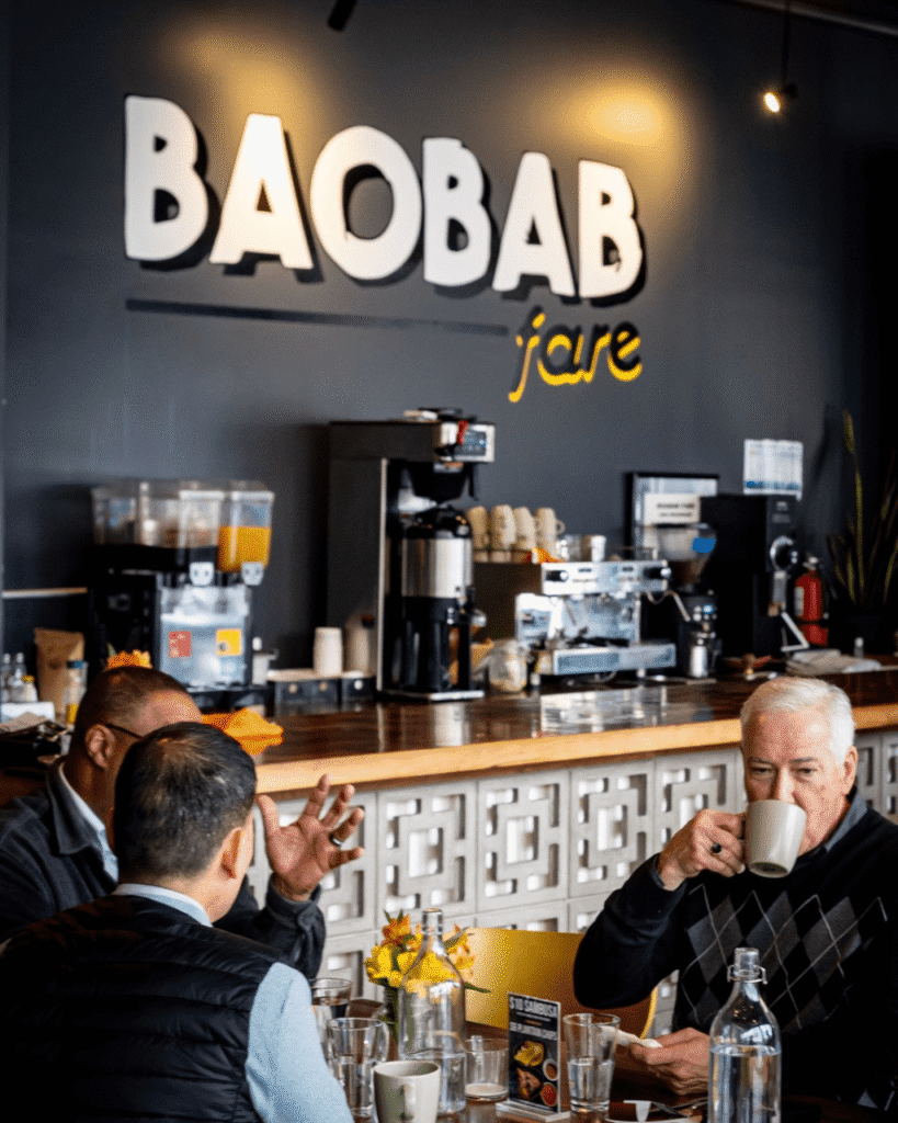 Three people sitting at a table at the Baobab Fare restaurant. One person is talking, another person is listening and the third person is drinking from a mug
